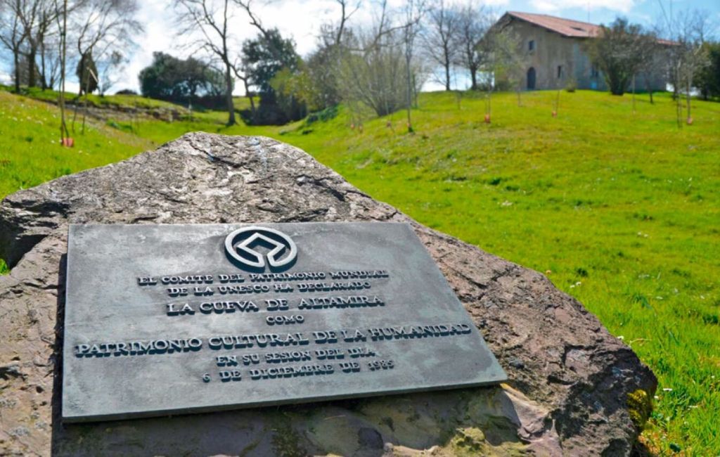 Placa en los alrededores de la cueva de Altamira que recuerda su declarción como Patrimonio Cultural de la Humanidad de la UNESCO en el año 1985. La entrada de la cueva está al fondo.